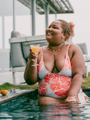 Femme ronde souriante portant un maillot une pièce imprimé corail avec un verre de jus à la main. 